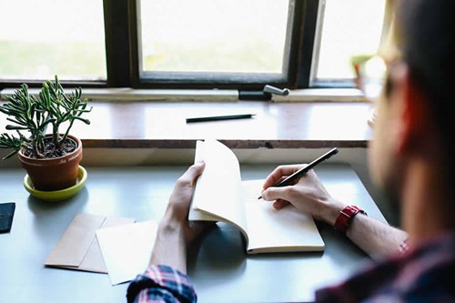 man with glasses writing in journal with house plant on desk