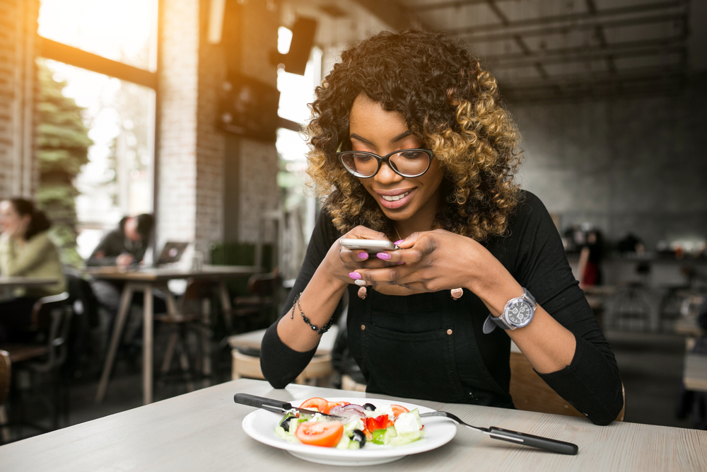 dark-skinned woman with curly hair taking photo of meal at restaurant